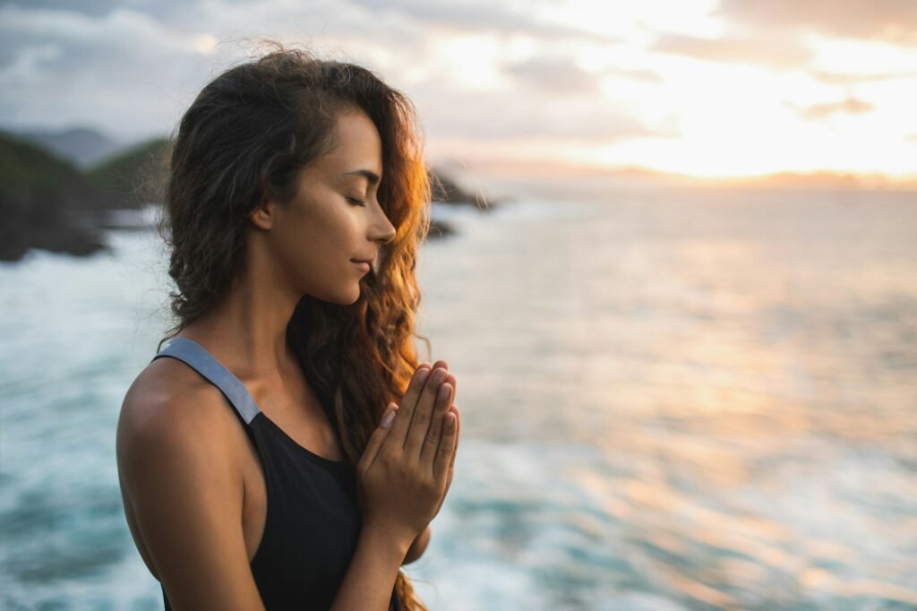 Young woman praying and meditating alone at sunset with beautiful ocean and mountain view