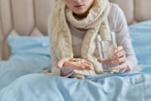 Female taking pills, capsules with glass of water, at home in bed