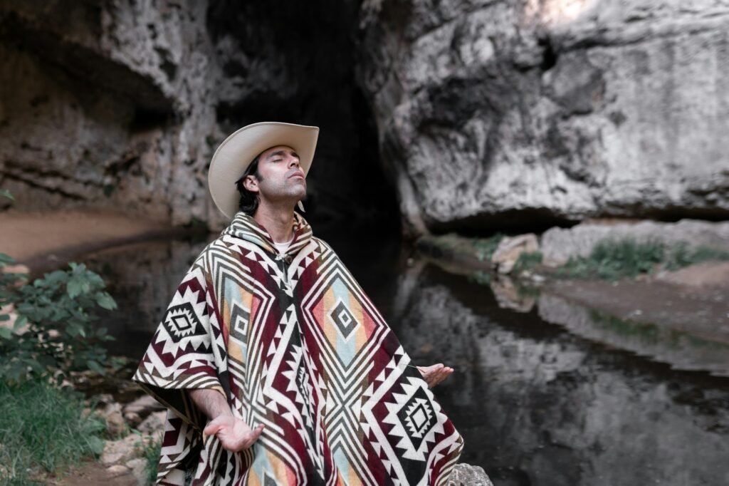 A mexican man wearing a poncho and a hat is meditating in the mountains