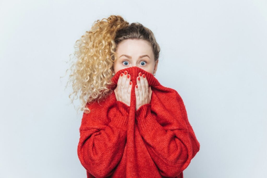 Frightened stunned female sees her phobia, covers face with collar of red sweater